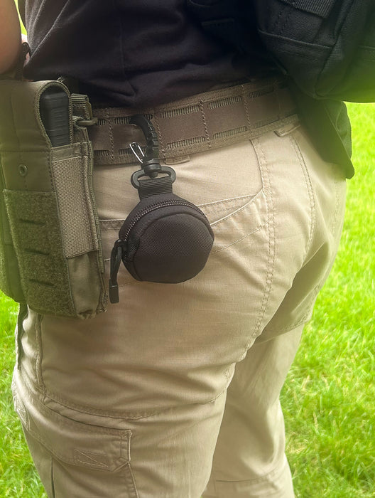 Close-up of a person wearing tactical gear with a small, round black device clipped to their belt, suitable for outdoor or tactical use.